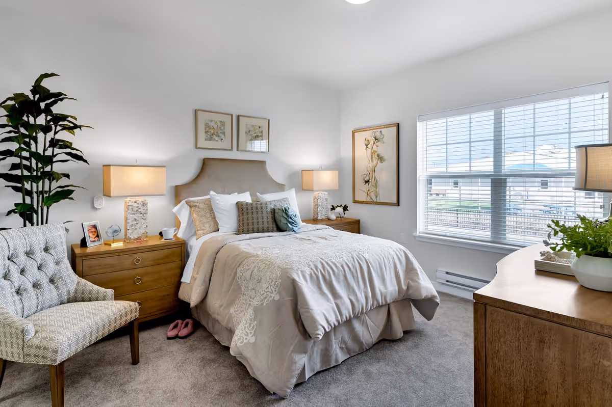 Bright, tastefully decorated bedroom with a bed dressed in neutral linens, two nightstands with lamps, an armchair, dresser, and a large window.