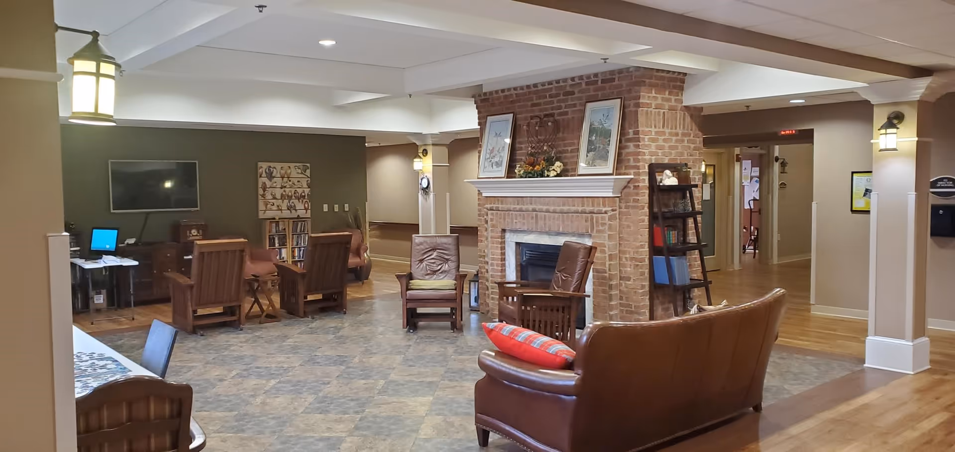 A cozy common area in a retirement and assisted living facility featuring a brick fireplace with framed artwork and floral arrangement on the mantel. Surrounding the fireplace are several wooden and leather chairs, a leather couch with a red plaid pillow, and a small bookshelf. The room has a mix of tiled and wooden flooring, warm lighting fixtures, and a television mounted on a green accent wall in the background.