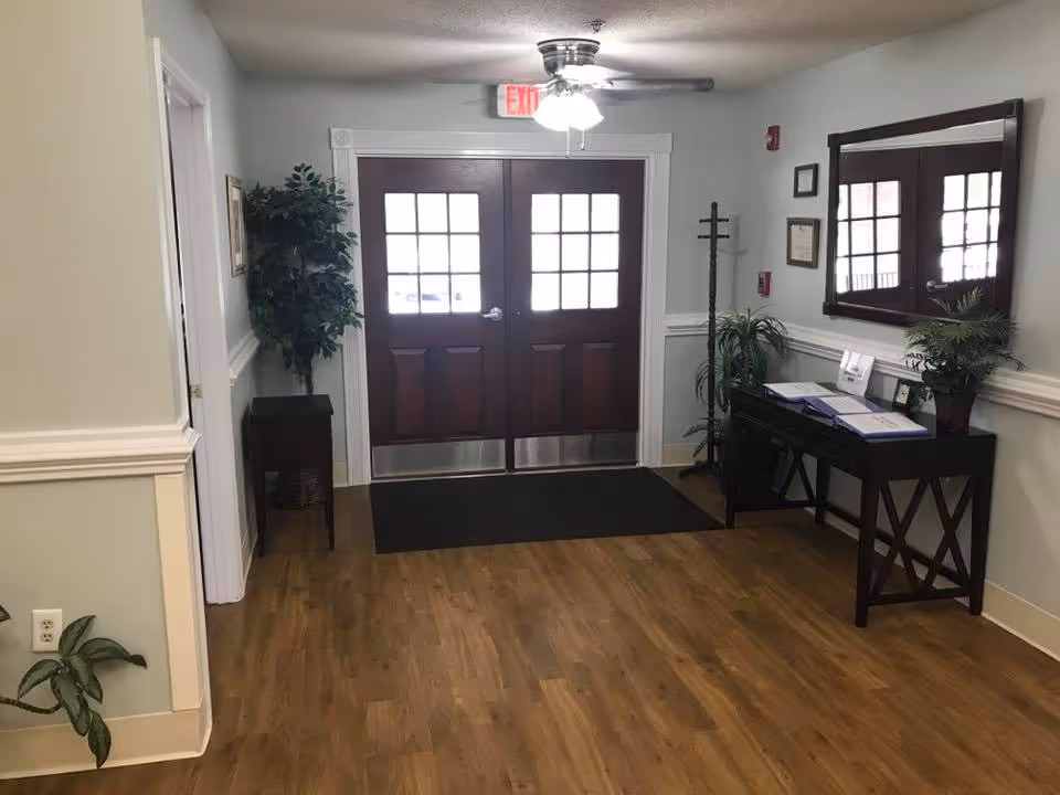 Interior view of an entrance area with double wooden doors featuring glass panes. The space has light-colored walls with white trim, wood flooring, a ceiling fan with lights, a black mat in front of the doors, a small table with plants and documents on the right side, a coat rack, and a large mirror above the table.