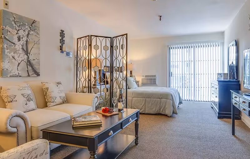 A cozy senior living room and bedroom area with a beige sofa adorned with butterfly-patterned pillows, a black coffee table with books and a wine bottle, a decorative folding screen separating the living space from the bedroom, a neatly made bed with white bedding, a dresser, and vertical blinds covering a large window or sliding door.