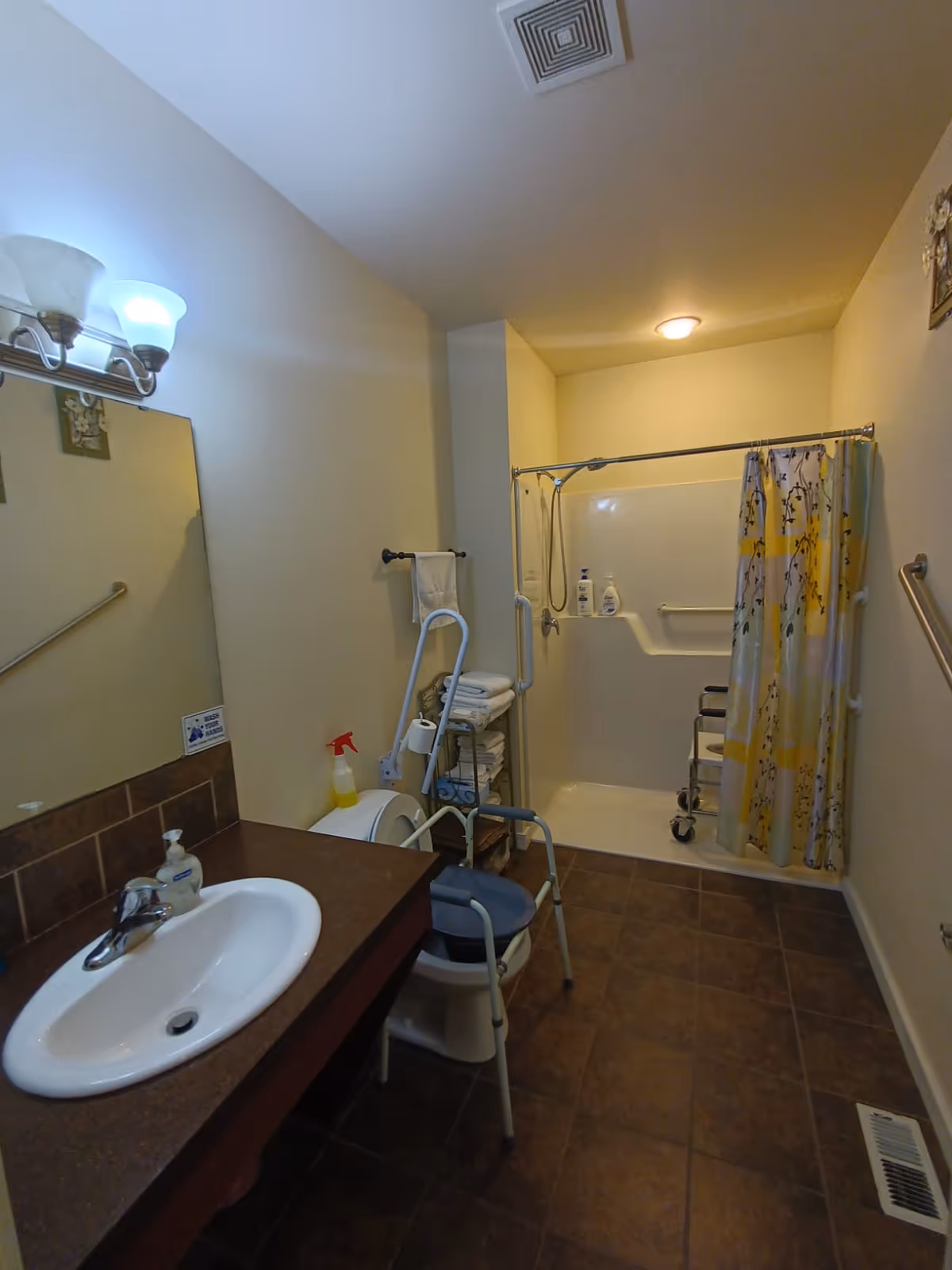 A bathroom with a sink and mirror on the left, a toilet with a raised seat and safety frame, a metal rack with folded towels, and a walk-in shower with a floral shower curtain and grab bars. The floor is tiled and the walls are painted light yellow.