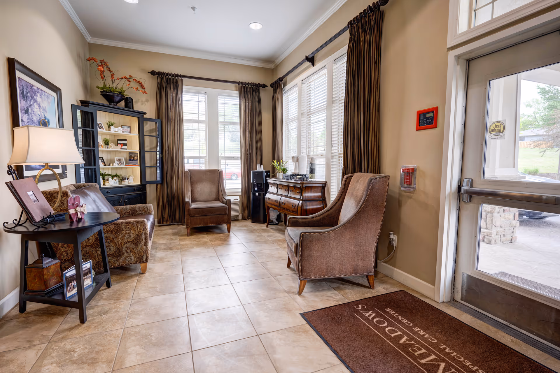 A cozy sitting area in a senior living facility with two upholstered armchairs, a small side table with a lamp, a black cabinet with shelves displaying decorative items, and large windows with brown curtains letting in natural light. There is a glass door on the right side leading outside, and a brown floor mat near the entrance.