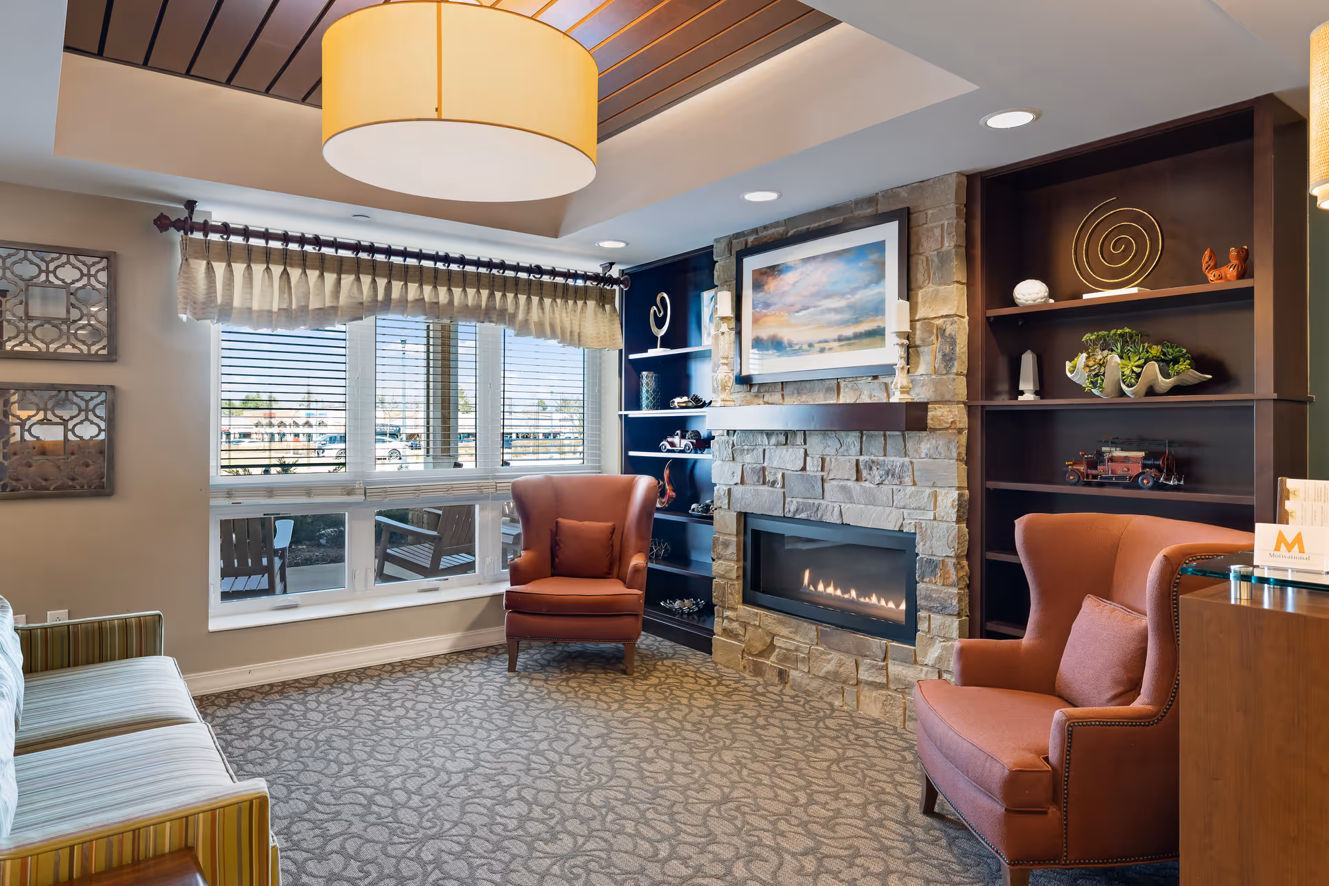 A cozy living room area with two orange armchairs and a striped sofa. There is a stone fireplace with a fire burning and a framed landscape painting above it. Built-in dark wood shelves flank the fireplace, decorated with various ornaments and plants. A large window with blinds and a valance lets in natural light, and a modern ceiling light fixture hangs above.