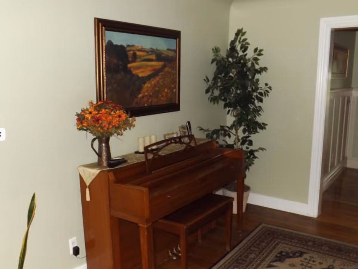 A wooden piano with a matching bench is placed against a light green wall. On top of the piano, there is a vase with orange flowers, three candles, and a few small decorative items. Above the piano hangs a framed landscape painting. To the right of the piano is a tall potted plant. The floor is wooden with a patterned rug partially visible. An open doorway leads to another room with white wainscoting.