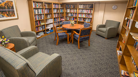 Carpeted community library with shelves of books, a round wooden table surrounded by chairs, and several upholstered armchairs.