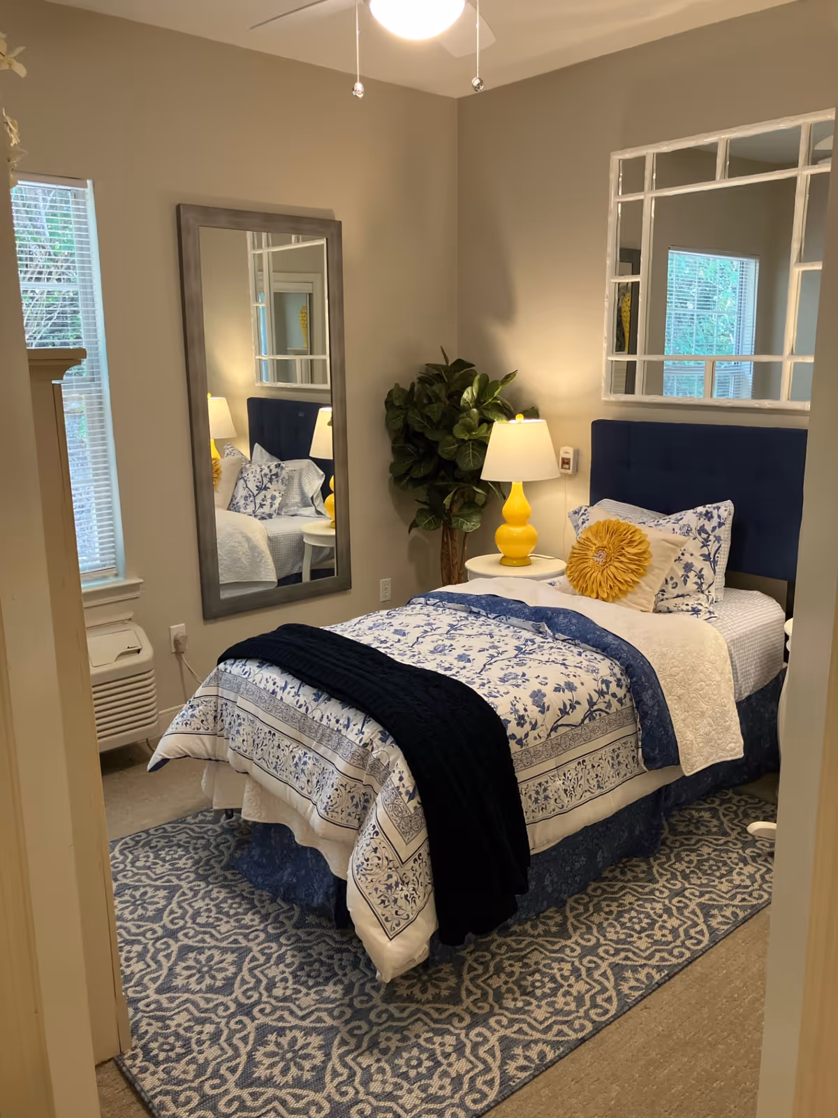 A cozy bedroom with a single bed dressed in blue and white floral bedding, a dark blue throw blanket, and a yellow decorative pillow. There is a white nightstand with a yellow lamp next to the bed. A large rectangular mirror hangs on the wall beside a window with blinds. A green potted plant is placed in the corner, and a patterned blue and white rug covers the floor.