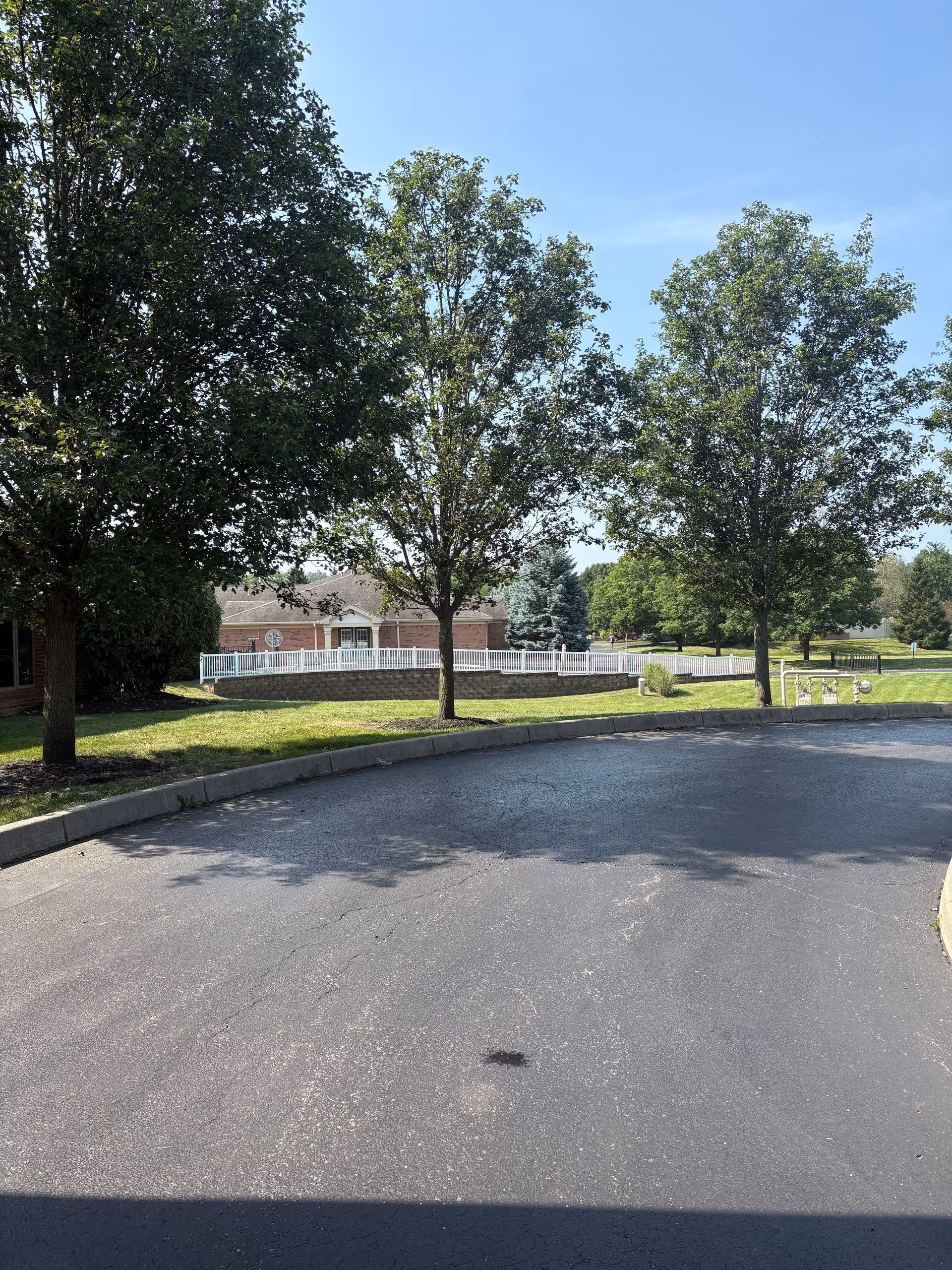 A paved driveway curves around a grassy area with three leafy trees. In the background, there is a brick building with a white fence and more trees under a clear blue sky.