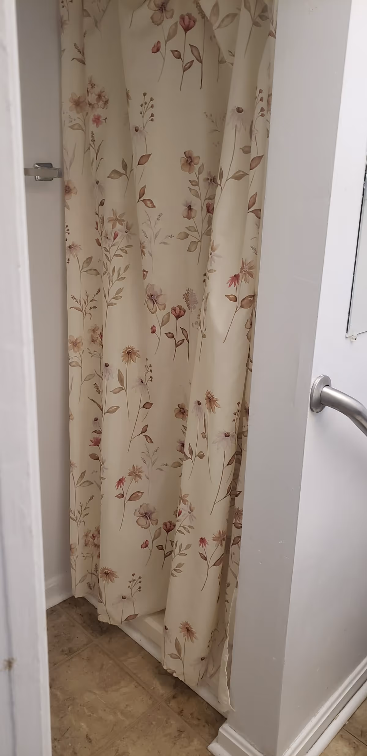 A small shower area with a floral patterned shower curtain in soft beige tones. The shower is enclosed by white walls, and a silver door handle is visible on the right side. The floor outside the shower has beige tiles.