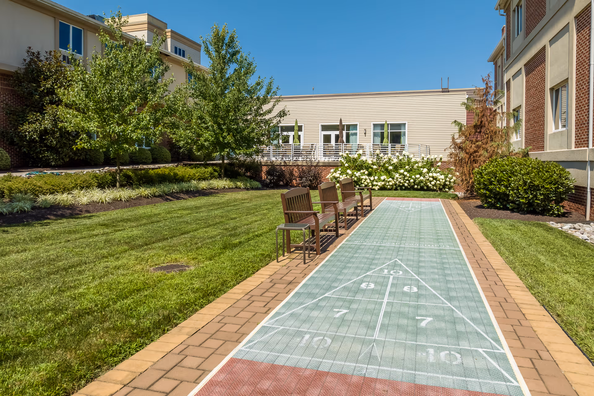 Outdoor shuffleboard court with four wooden chairs lined up along one side, surrounded by well-maintained grass, bushes, and trees. The court is situated between two buildings under a clear blue sky.