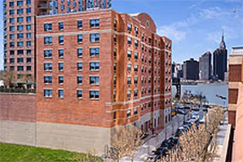 Exterior view of a large red brick riverside building with parked cars along the street.
