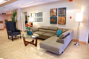 A bright lounge area with an L-shaped gray sofa, armchair, coffee table, floor lamp, potted plants and wall art in a senior facility.