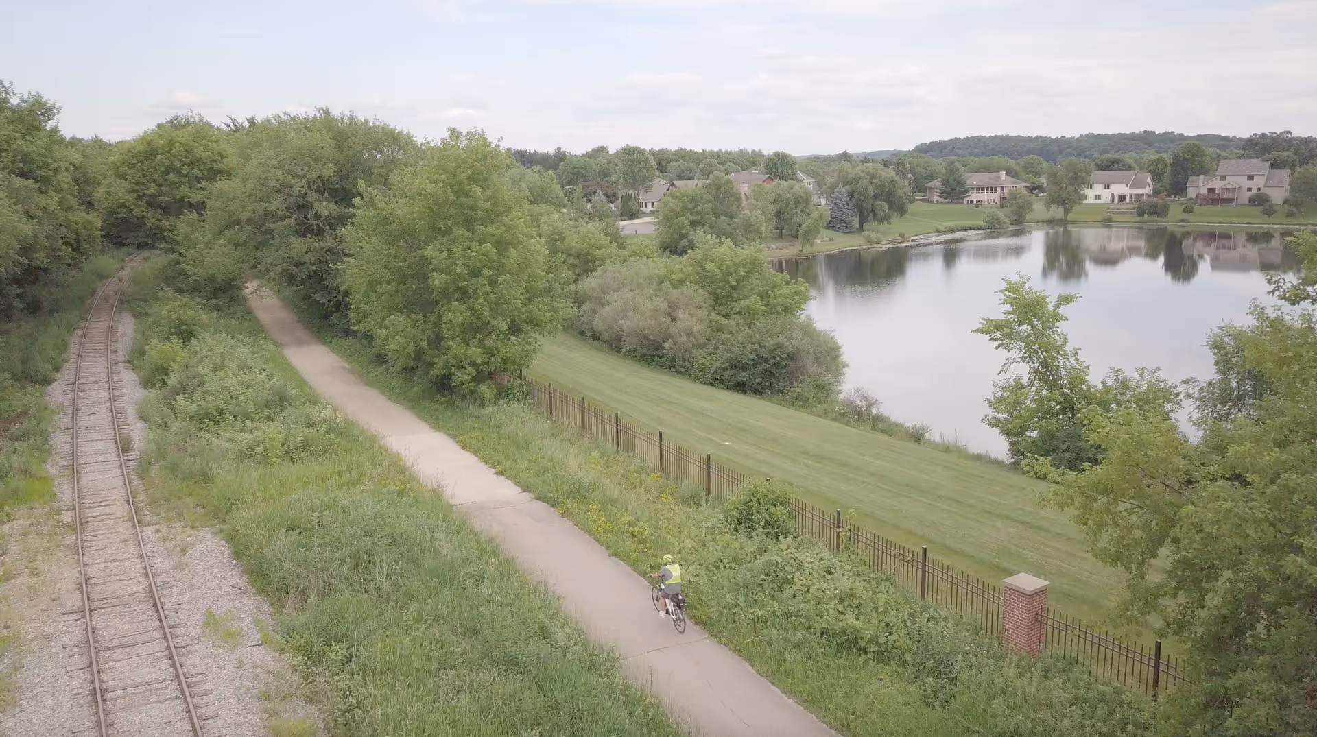 A scenic outdoor view showing a cyclist riding on a paved path next to a railway track. The path is bordered by lush green trees and bushes, with a calm lake and houses visible in the background under a partly cloudy sky.