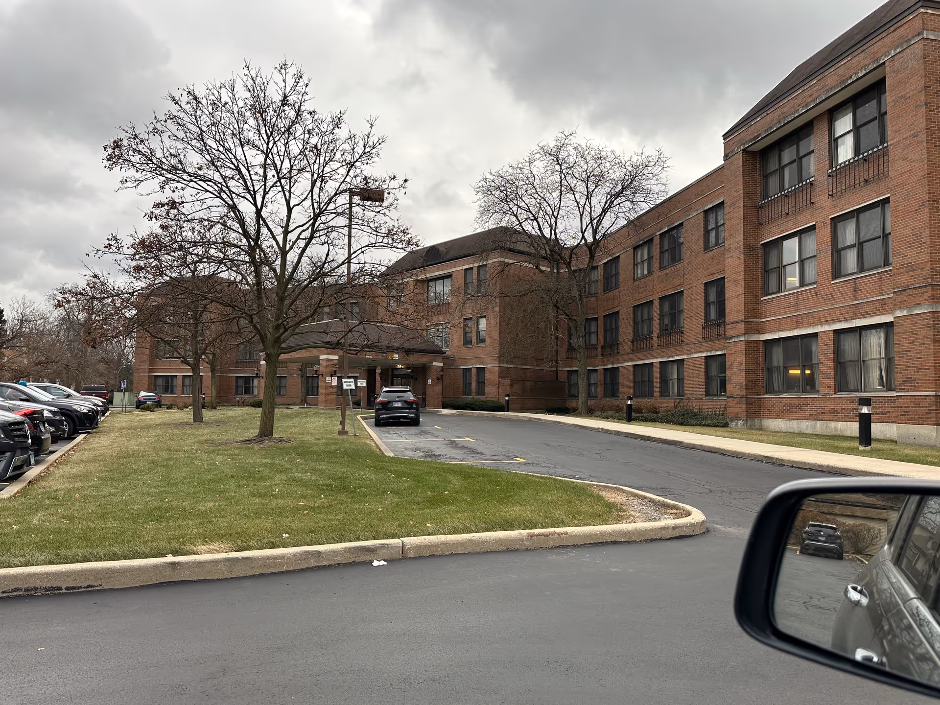 Exterior view of a three-story brick nursing home building with multiple windows, a driveway leading to a covered entrance, several leafless trees, a grassy area, and parked cars along the side. The sky is overcast and a car's side mirror is visible in the foreground.