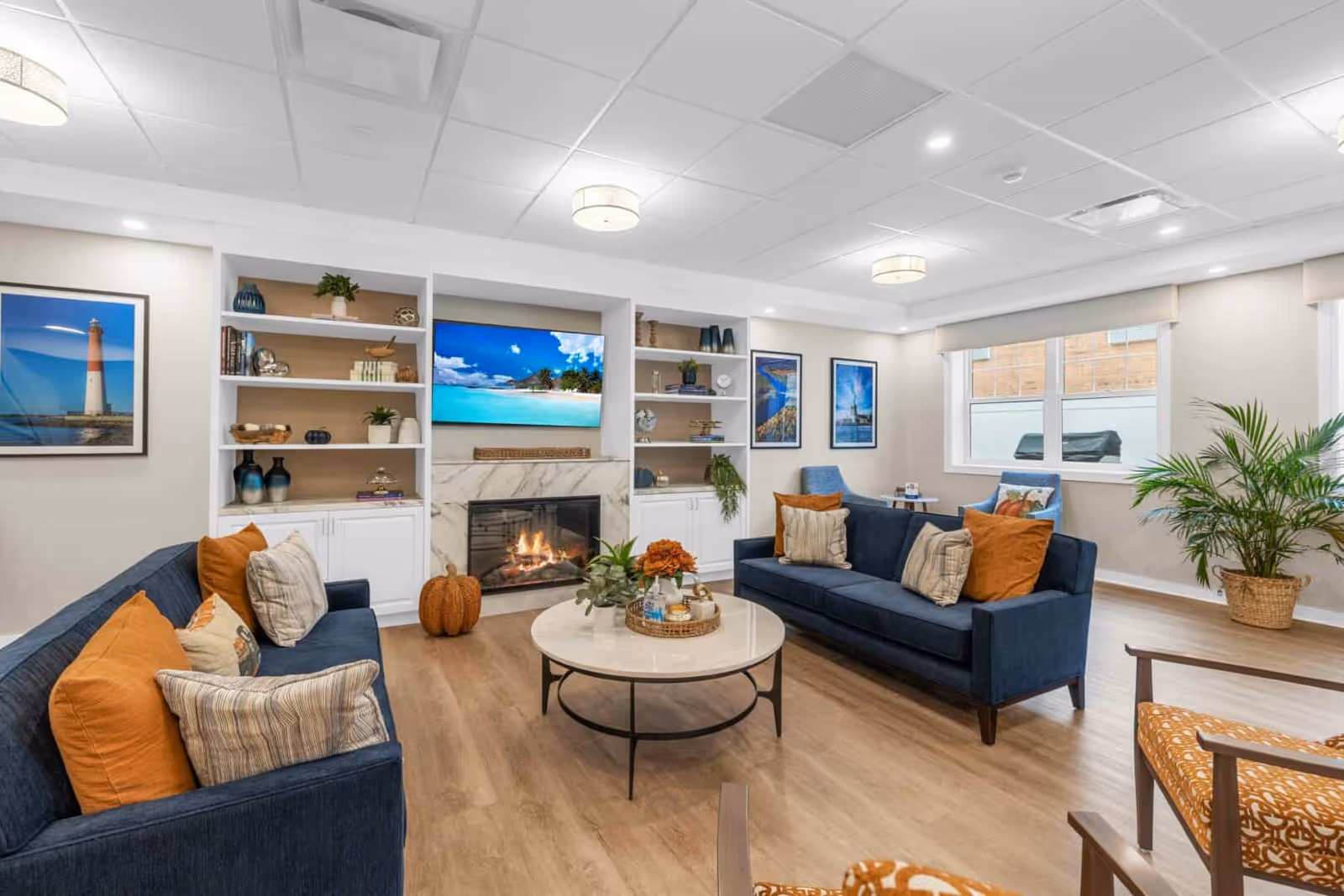 A bright and modern living room with two navy blue sofas adorned with orange and beige pillows, a round white coffee table with decorative items, built-in white shelves with books and plants, a fireplace with a marble surround, a flat-screen TV displaying a beach scene, framed pictures on the walls, large windows, and a potted plant in the corner.