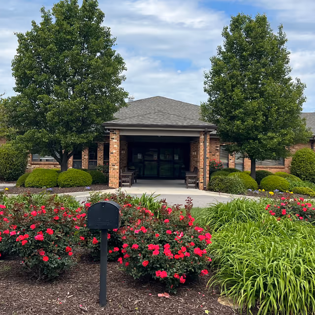 Front entrance of a brick building with a covered porch, flanked by two large trees and well-maintained landscaping including bushes and red flowers in the foreground under a partly cloudy sky.