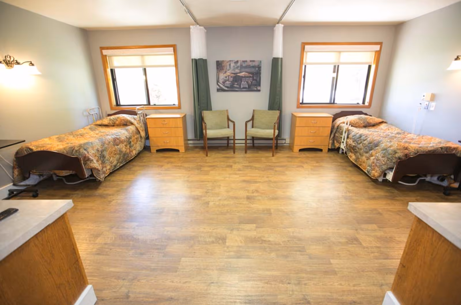 A shared bedroom in a senior living facility with two single beds on opposite sides, each with floral bedspreads. Between the beds are two wooden nightstands and two green cushioned chairs placed against the wall. Two windows with blinds allow natural light into the room, and a painting hangs on the wall between the windows. The floor is wood, and the room is well-lit with wall-mounted lamps.