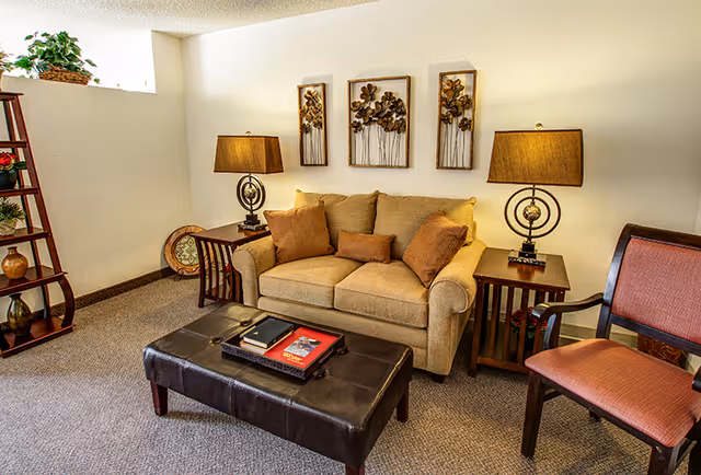 A cozy living room area with a beige loveseat adorned with brown cushions, flanked by two wooden side tables each holding a lamp with a beige shade. Above the loveseat are three decorative wall art pieces featuring floral designs. In front of the loveseat is a dark brown leather ottoman with books and a tray on top. To the right is a wooden chair with a reddish cushion, and to the left is a wooden shelf with decorative items and a plant on top. The room has beige carpet and white walls with a small window near the ceiling.