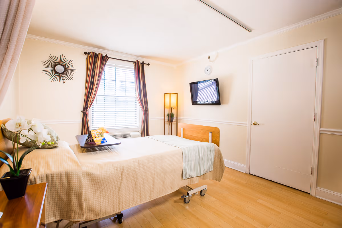A bright and clean bedroom in a senior living facility featuring a single bed with beige bedding, a small wooden side table with a potted plant, a window with striped curtains, a wall-mounted TV, a standing lamp, and a closed white door.