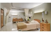 A clean and well-lit hospital-style room with a single bed covered in beige linens. The room features a wooden nightstand with decorative giraffe figurines, a wooden dresser, a wall-mounted light, and privacy curtains. The walls are painted light green, and the floor is white and shiny.