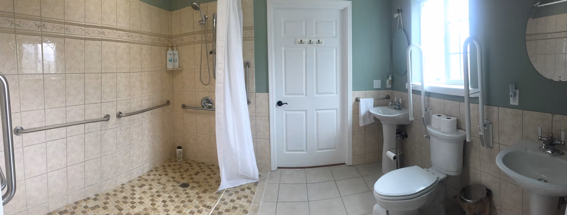 A spacious, accessible bathroom with beige tiled walls and floor. The bathroom features a walk-in shower with a white curtain, grab bars on the walls, a white door, a pedestal sink, a toilet with grab bars, a round mirror, and a window letting in natural light.