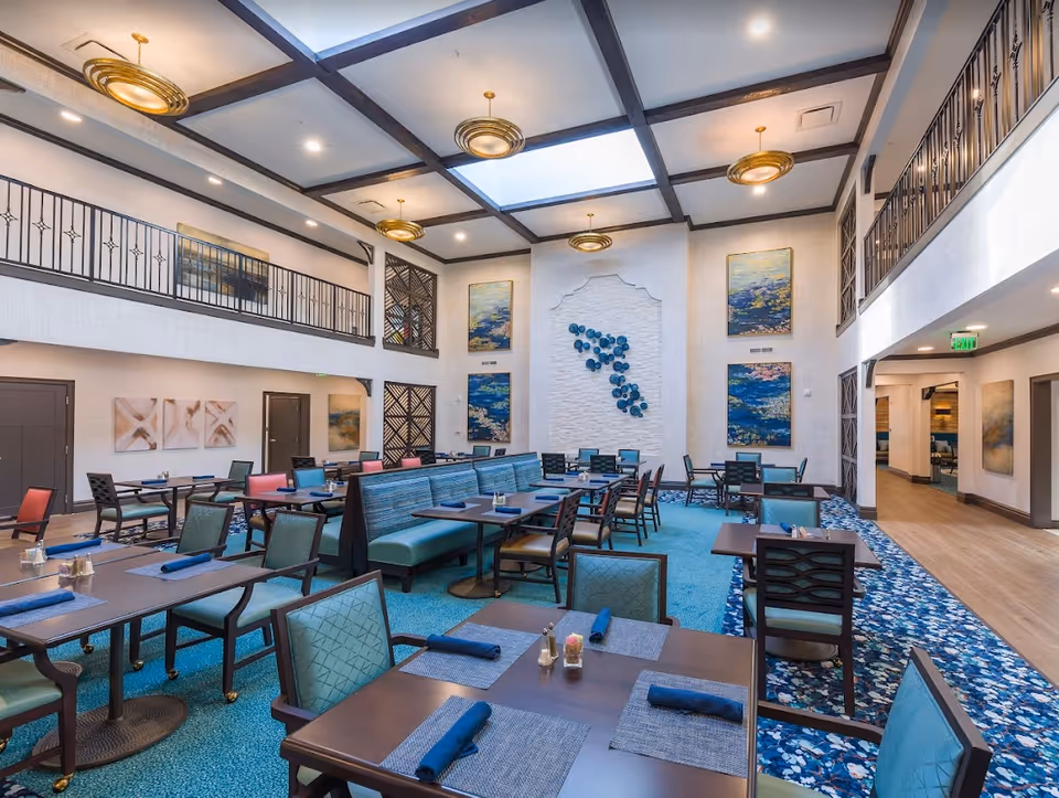 A spacious, well-lit dining room with high ceilings and skylights. The room features multiple tables set with blue napkins and placemats, surrounded by chairs with green upholstery. There is a long booth seating area in the center. The walls are decorated with large abstract paintings and a decorative wall installation. The room has a blue and patterned carpet, and there is a balcony railing on the upper level.