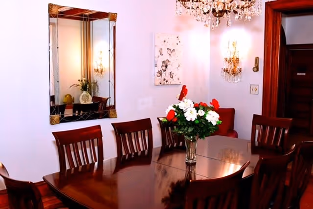 A dining room with a polished wooden table surrounded by wooden chairs. A vase with white and red flowers is placed in the center of the table. The room features a large mirror on the wall, a chandelier hanging from the ceiling, wall sconces, and a piece of artwork on the wall. There is a wooden door visible in the background.