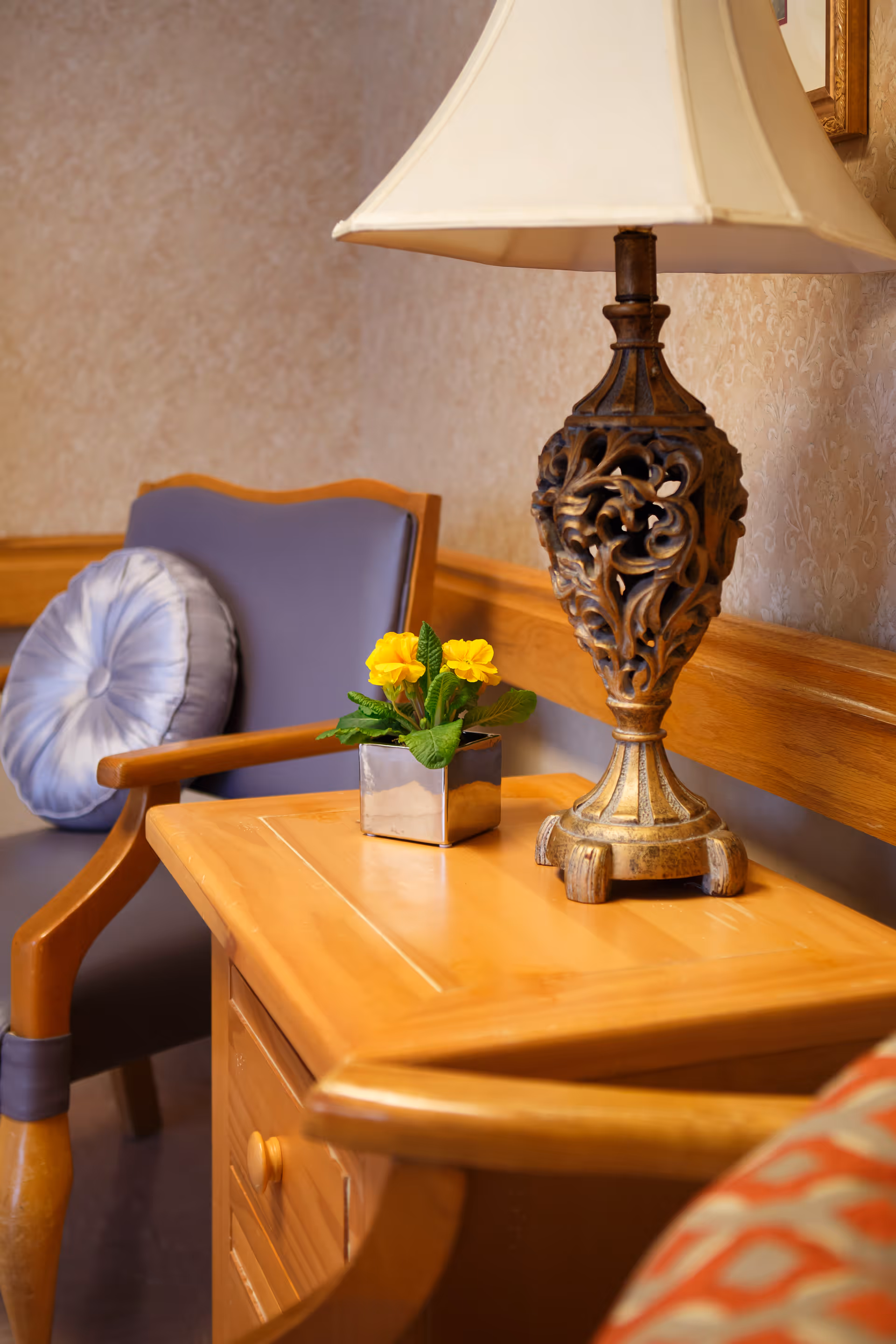 A cozy corner with a wooden side table holding a decorative lamp with an ornate base and a small potted plant with yellow flowers. Next to the table is a wooden armchair with a purple cushion and a round, pleated pillow. The background features a beige patterned wallpaper and a wooden chair rail.