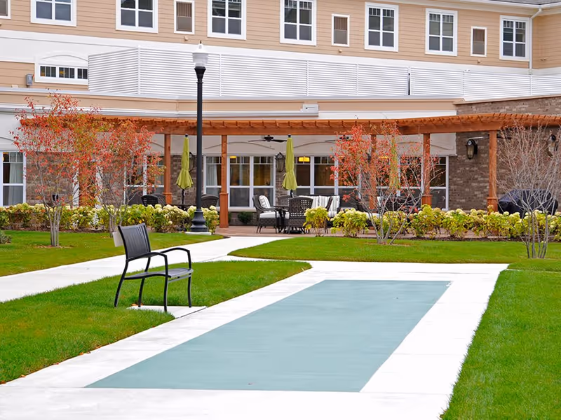 Outdoor courtyard of a senior living facility with a pergola-covered seating area, walkways, a bench and manicured lawns.