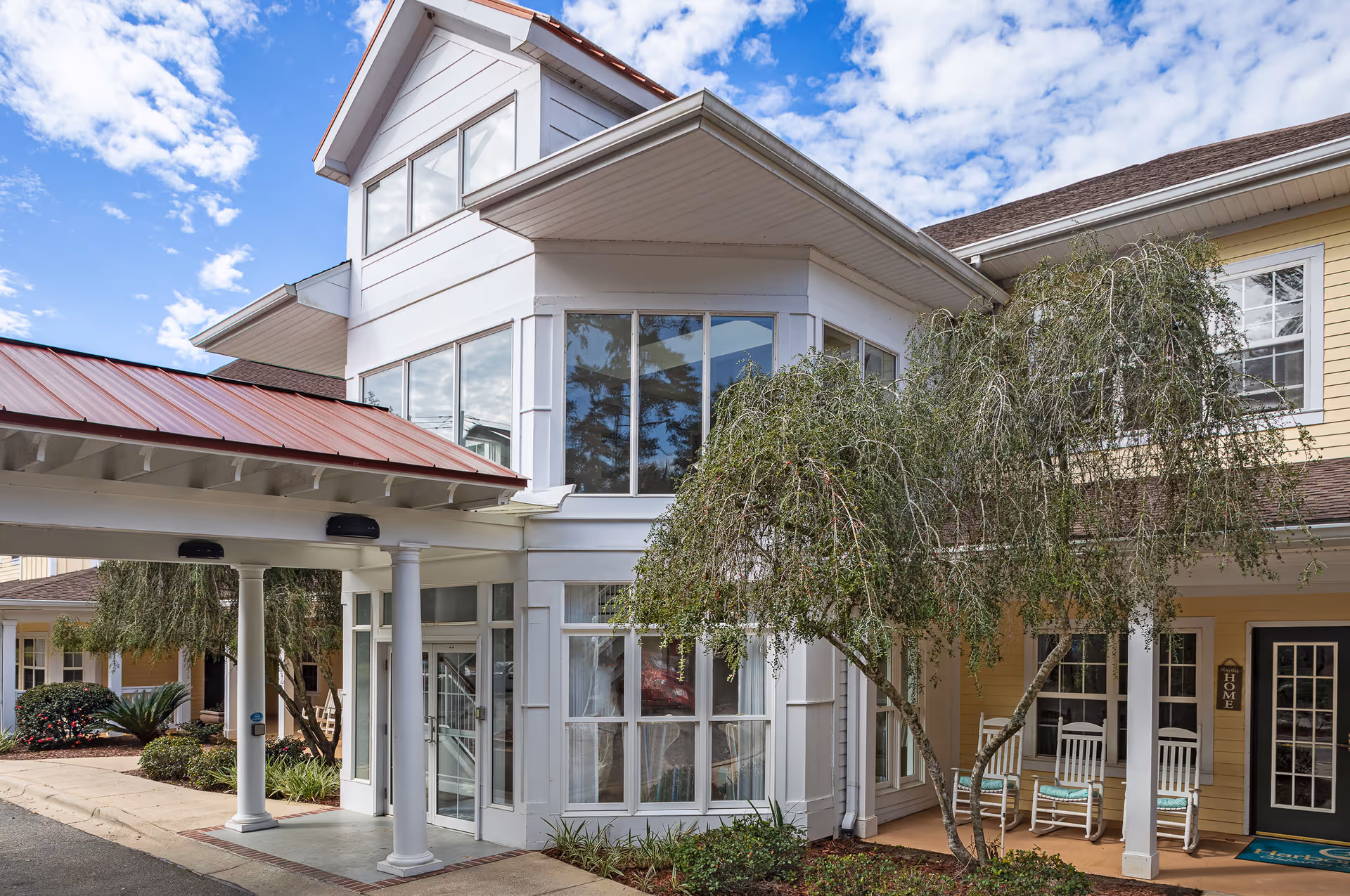 Exterior view of The Gables of Tallahassee senior living facility entrance with white columns, large windows, a covered drop-off area with a red roof, and a small tree in front. There are rocking chairs on the porch and a blue welcome mat near the door.