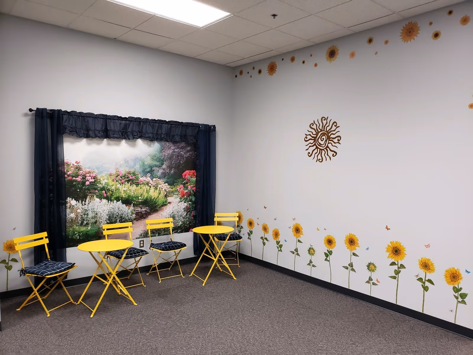 A corner of a room with two small yellow tables and four yellow chairs with dark cushions. The walls are decorated with sunflower and butterfly decals, and a large mural of a garden path with flowers is framed by black curtains. The ceiling has white tiles and the floor is carpeted in gray.