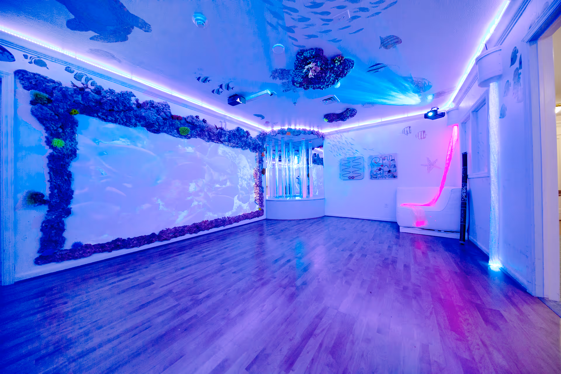 A sensory room with an underwater theme featuring blue and purple lighting. The walls and ceiling are decorated with images of fish and coral reefs. There is a large illuminated panel on one wall resembling an aquarium, a bubble tube in the corner, and a white cushioned chair with a pink glowing strip of light. The room has wooden flooring and a calm, immersive atmosphere.