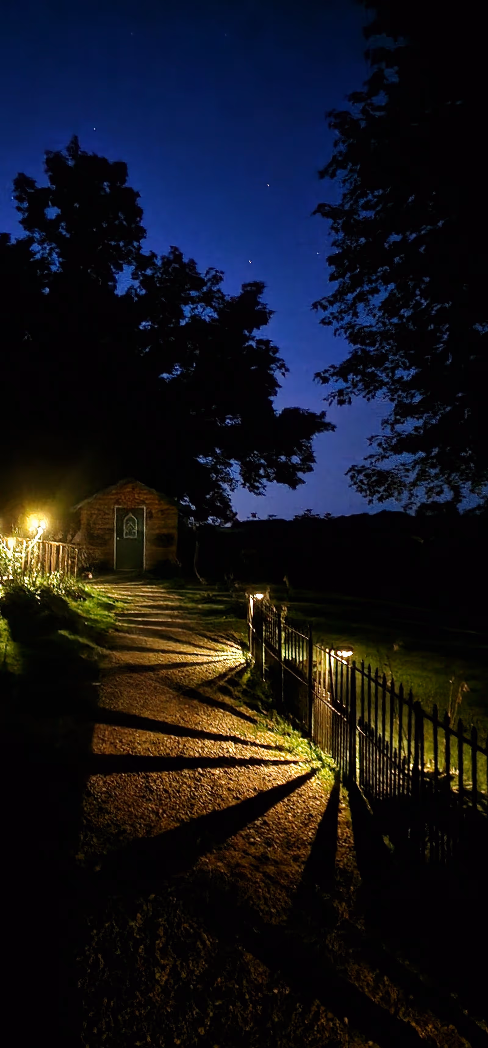 A nighttime outdoor scene showing a gravel pathway lit by small ground lights casting long shadows of a metal fence. The path leads to a small building with a door, surrounded by large trees under a dark blue sky with visible stars.