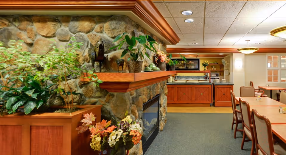 Cozy dining area with a stone fireplace, plants, a buffet counter, and tables with chairs.