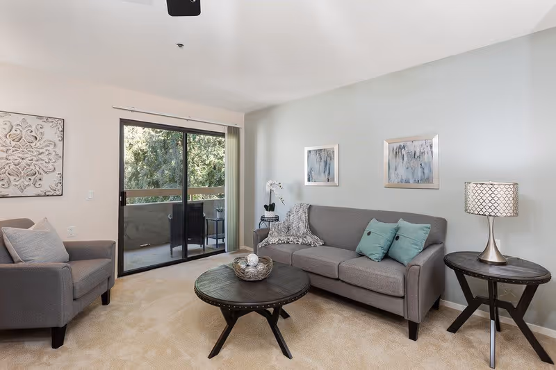 A cozy living room with a gray sofa adorned with two teal pillows and a gray throw blanket, a matching gray armchair with a light gray pillow, a round wooden coffee table with decorative items, and a round side table with a lamp. The room has light beige carpet, light blue and white walls with framed artwork, and a sliding glass door leading to a balcony with outdoor seating and greenery visible outside.