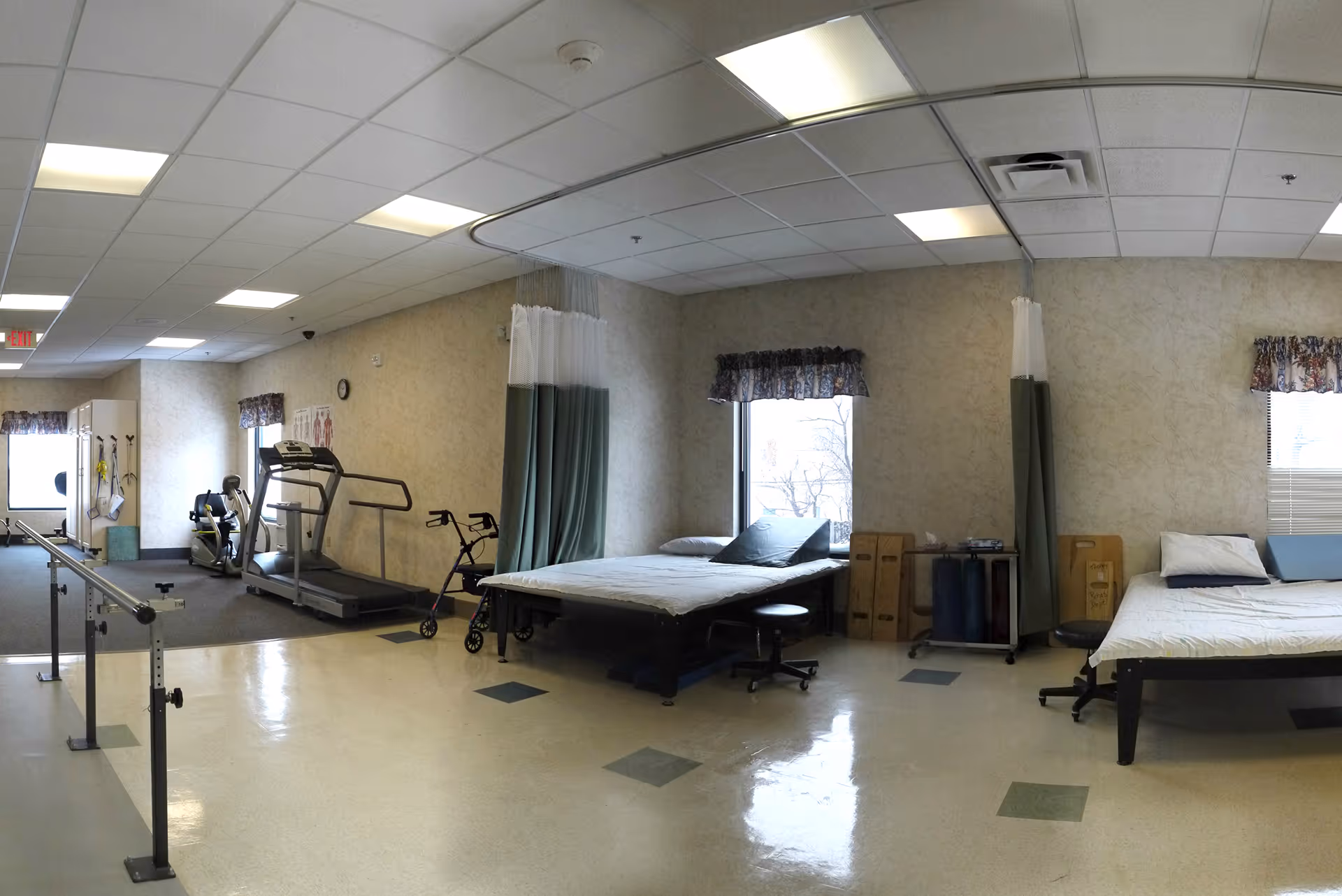 A rehabilitation or physical therapy room with two hospital beds separated by curtains, exercise equipment including a treadmill and stationary bike, parallel bars for walking practice, and various therapy tools. The room has large windows with floral valances and a tiled floor with some carpeted areas.