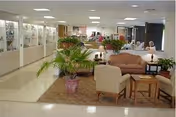 Interior view of a senior living facility lobby or common area with seating arrangements including a sofa, armchairs, and small tables. There are potted plants placed around the seating area, and display cases line the left wall. The space is well-lit with ceiling lights, and an American flag is visible in the background.