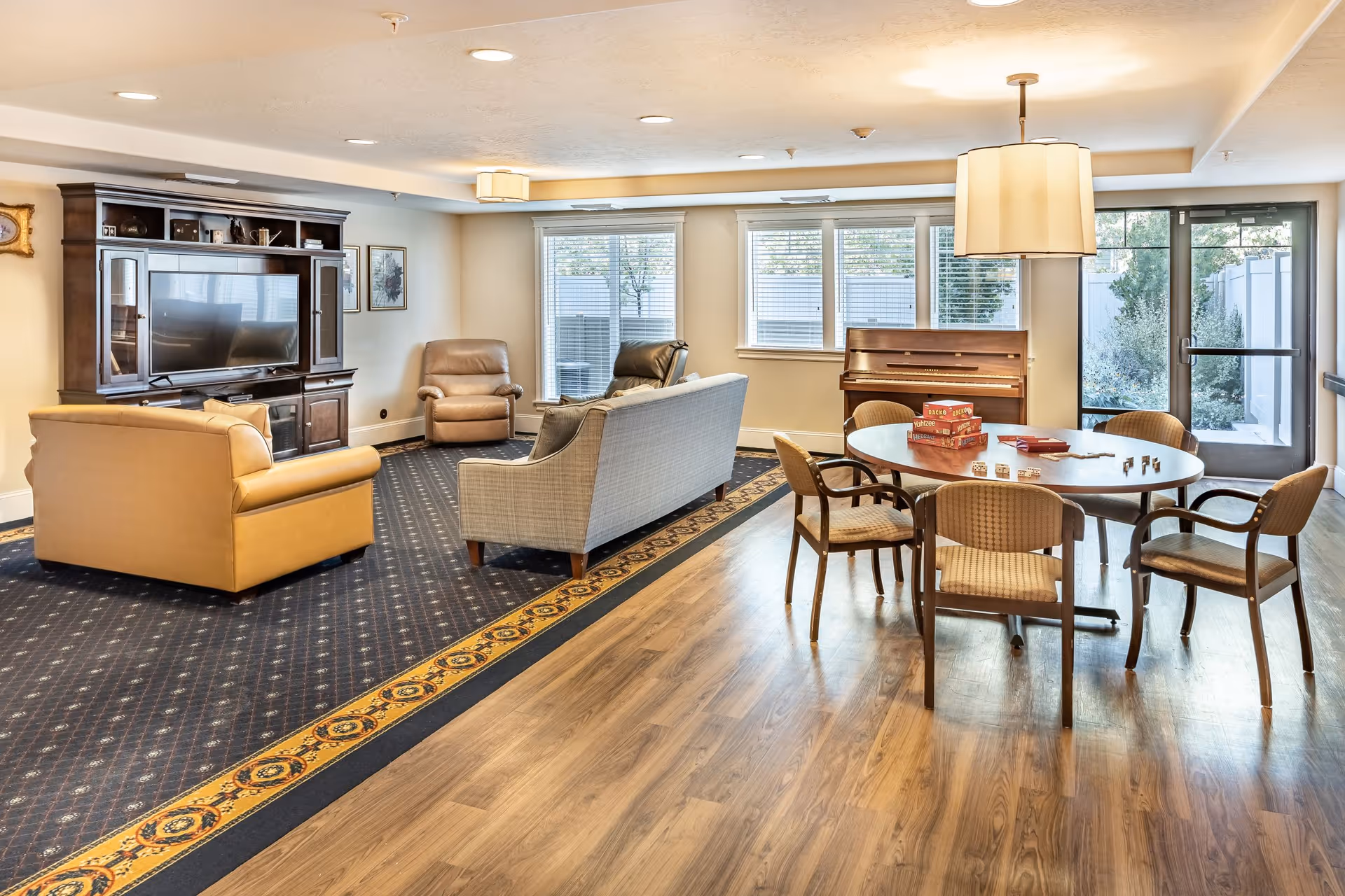 A bright and spacious common area in Legacy House of Spanish Fork featuring a seating area with a large TV, two armchairs, and a sofa on a patterned carpet. Adjacent to this is a wooden round table with six chairs, board games on the table, and a piano against the wall near large windows and a glass door leading outside.