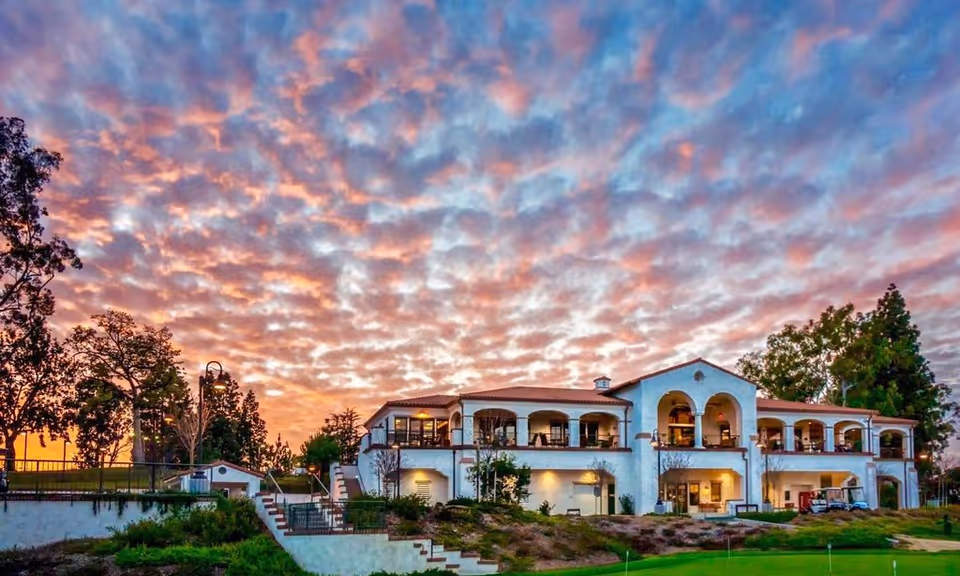 Two-story Mediterranean-style clubhouse and landscaped grounds under a dramatic colorful sunset sky.