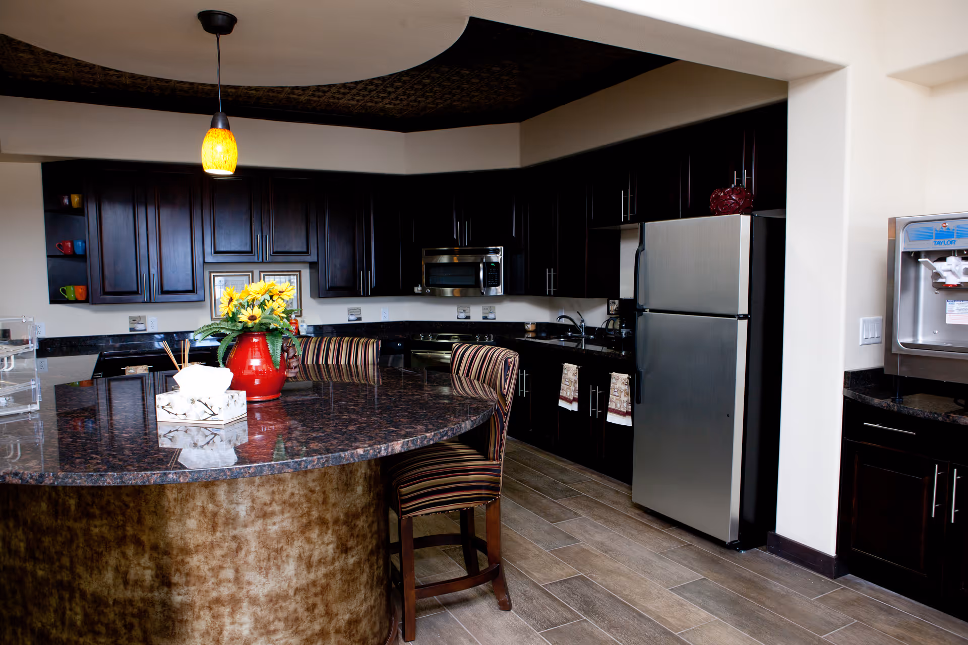 Modern communal kitchen with dark cabinetry, a large curved granite island topped with a red vase of sunflowers and striped chairs, and stainless steel appliances.