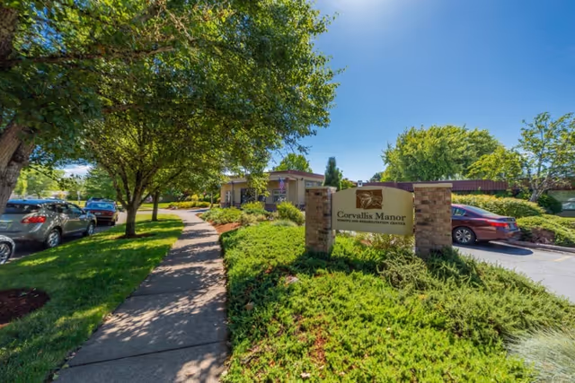 Exterior view of Corvallis Manor Nursing & Rehabilitation Center on a sunny day, showing a sidewalk lined with trees and parked cars, with the facility's sign prominently displayed in front of landscaped greenery.