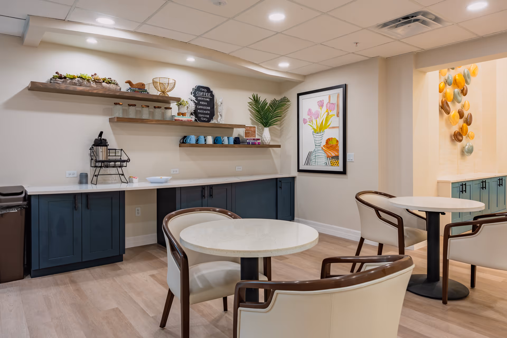 A cozy seating area with round tables and cushioned chairs in a senior living facility. The room features a countertop with cabinets underneath, floating shelves holding coffee mugs, jars, and decorative items. There is a coffee menu sign on the wall and framed artwork depicting flowers. The space is well-lit with recessed ceiling lights and has light wood flooring.