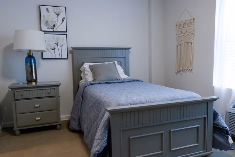 Neatly made single bed with a gray wooden frame and blue bedding in a bright, simply decorated bedroom with a nightstand and lamp.