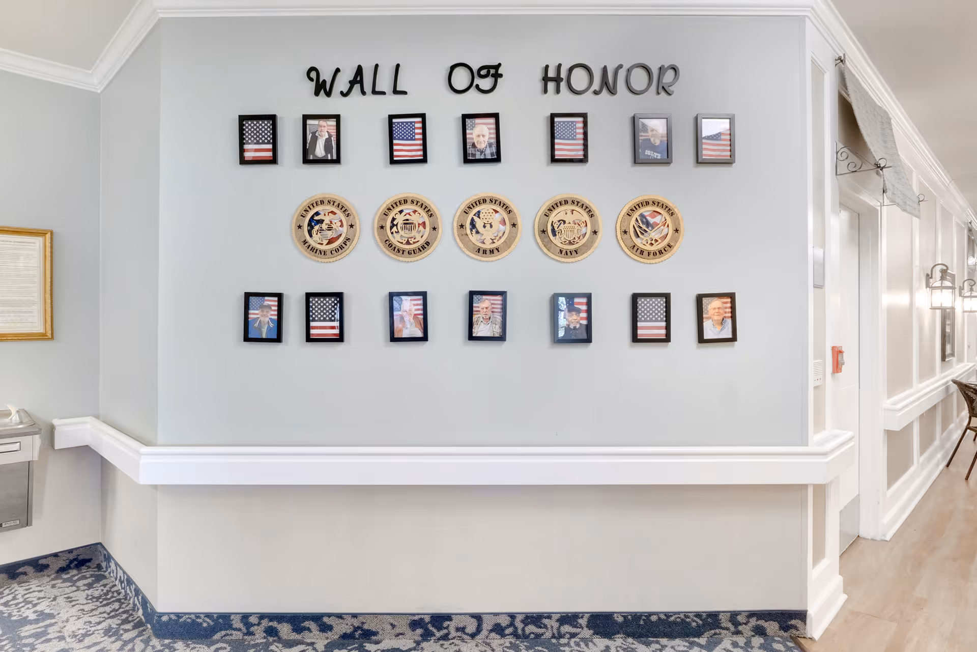 A hallway wall display titled 'Wall of Honor' featuring framed photos of veterans and American flags, along with emblems of the United States Marine Corps, Coast Guard, Army, Navy, and Air Force. The hallway has light-colored walls, a patterned carpet, and white trim with a water fountain and framed document on the left side.