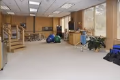 A spacious rehabilitation room with exercise equipment including therapy balls, parallel bars, and a set of stairs for physical therapy. The room has large windows with blinds, wood-paneled walls, and a light-colored carpeted floor. There are also plants and various therapy tools visible.