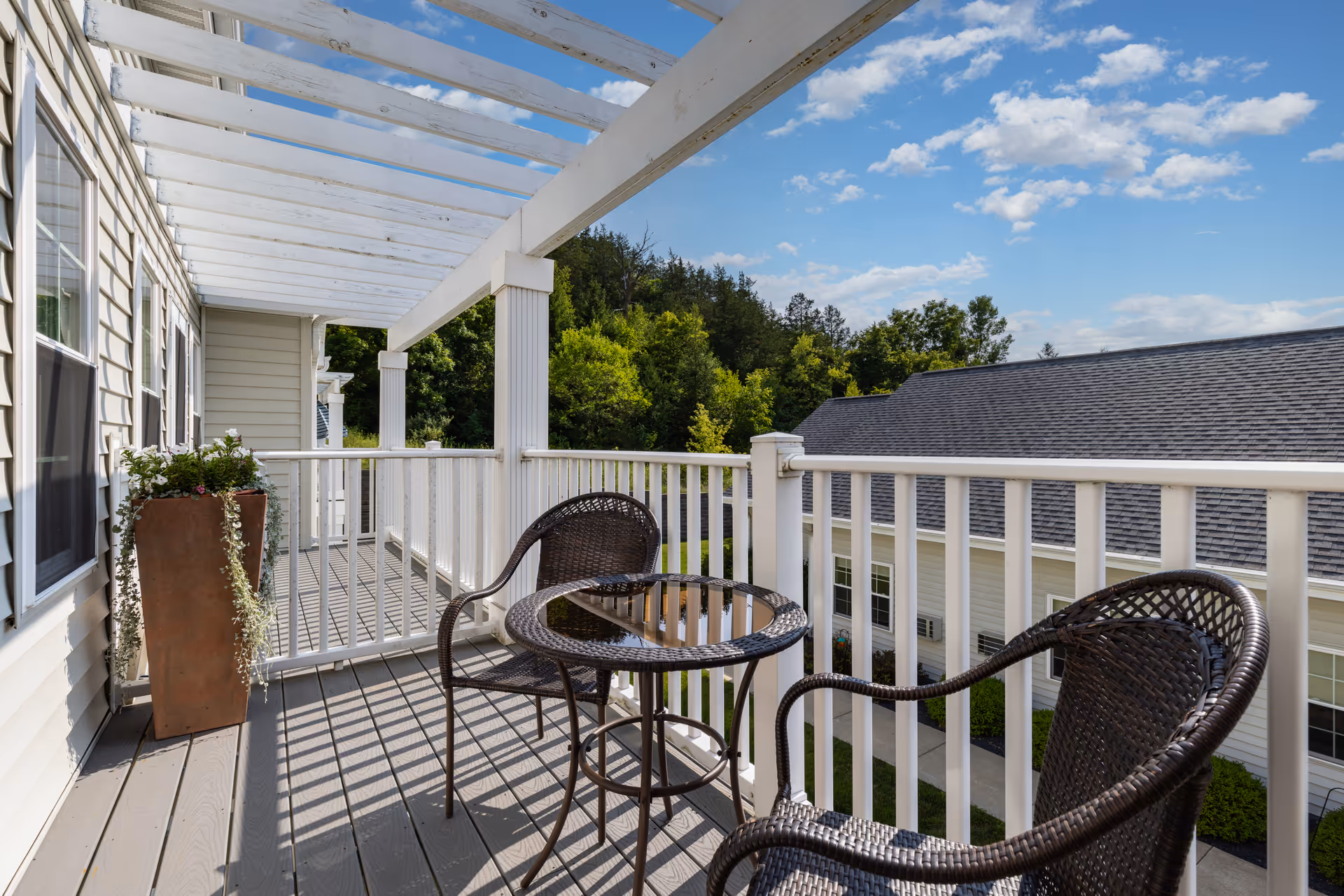 A balcony with white railings and a white pergola overhead, featuring two dark wicker chairs and a small round glass-top table. There is a tall planter with green plants on the left side. The balcony overlooks a building with a gray roof and white siding, with trees and a blue sky with scattered clouds in the background.