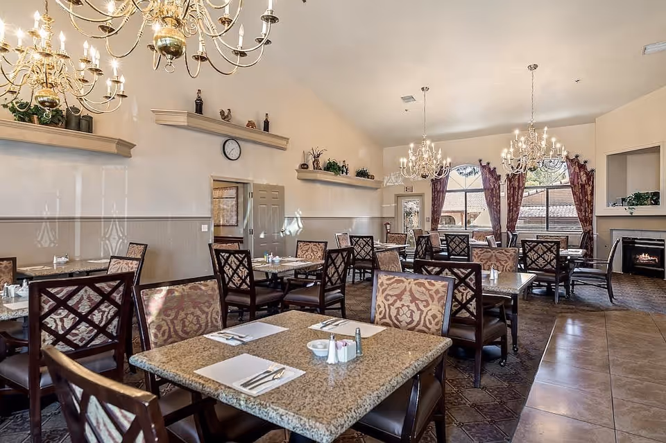 A spacious dining room with multiple tables and chairs arranged neatly. The tables have granite tops and are set with placemats, napkins, and cutlery. The room features elegant chandeliers hanging from the ceiling, large windows with patterned curtains allowing natural light to enter, and a fireplace in the corner. Shelves with decorative items and plants adorn the walls.