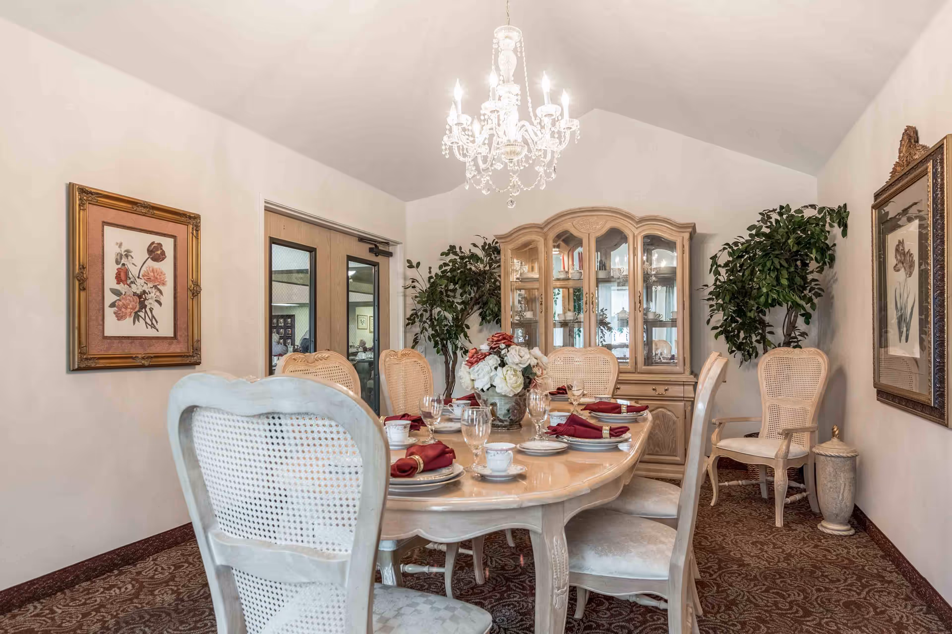 A formal dining room with a large wooden dining table set for six with plates, cups, glasses, and red napkins. The room features a chandelier hanging from the ceiling, a wooden china cabinet filled with glassware, two large potted plants, and framed floral artwork on the walls. The floor is carpeted with a patterned design.