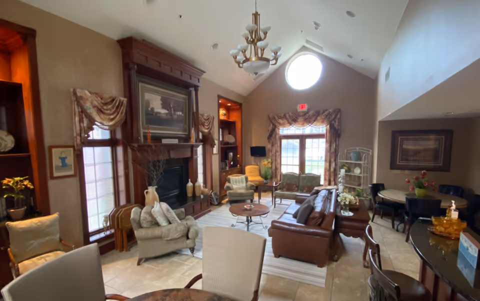 A spacious living room area with high vaulted ceilings and a large circular window near the ceiling. The room features a fireplace with a wooden mantel, flanked by two windows with draped curtains. There are several seating options including a leather sofa, cushioned armchairs, and a loveseat. A round coffee table is placed in the center on a striped area rug. In the background, there is a dining table with chairs and a shelving unit with decorative items. The room is warmly lit with a chandelier hanging from the ceiling.