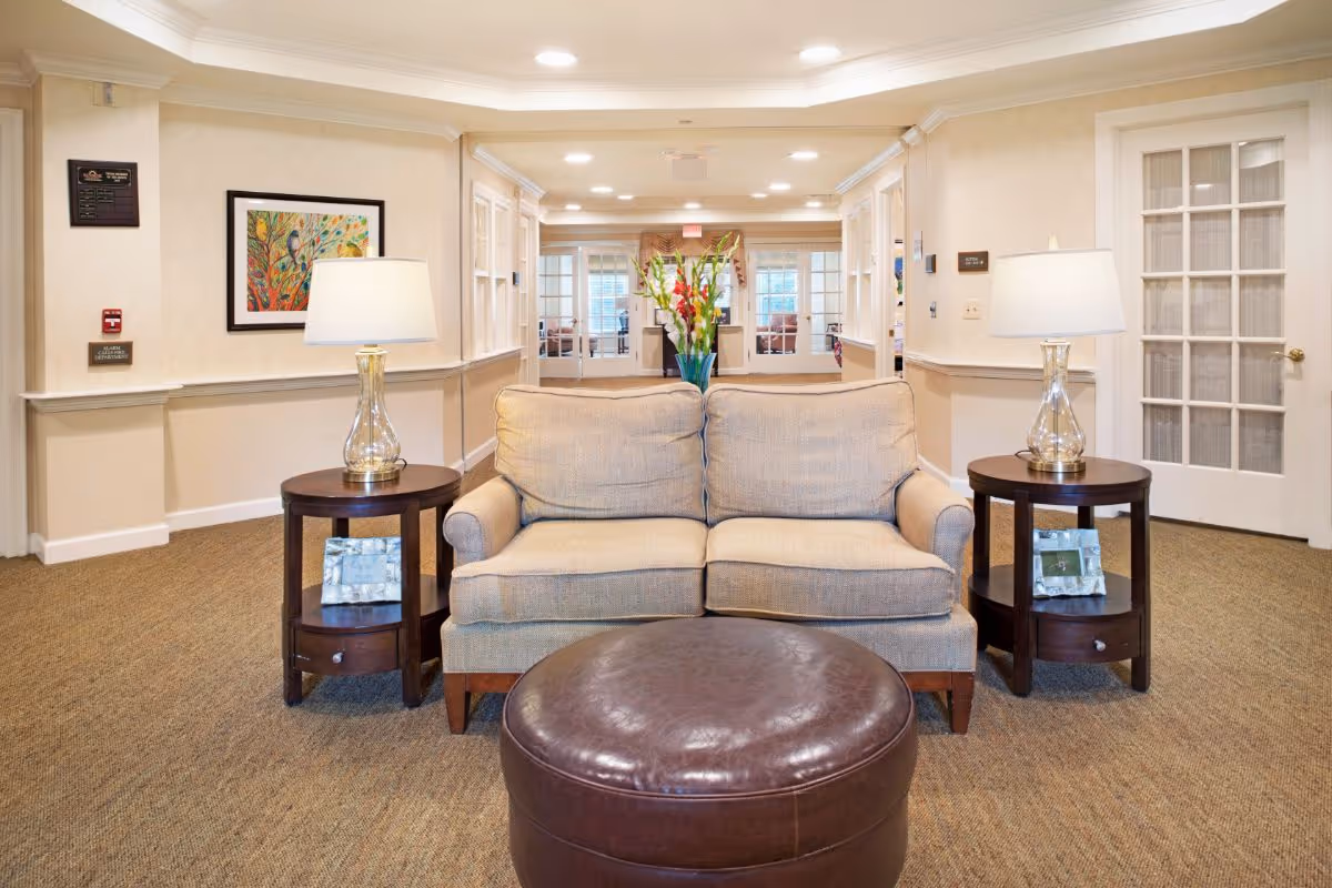 A cozy seating area in a senior living facility with a beige loveseat flanked by two round wooden side tables, each holding a glass lamp. In front of the loveseat is a round brown leather ottoman. The background shows a hallway with beige walls, framed artwork, and glass-paneled doors leading to another room with more seating and natural light.