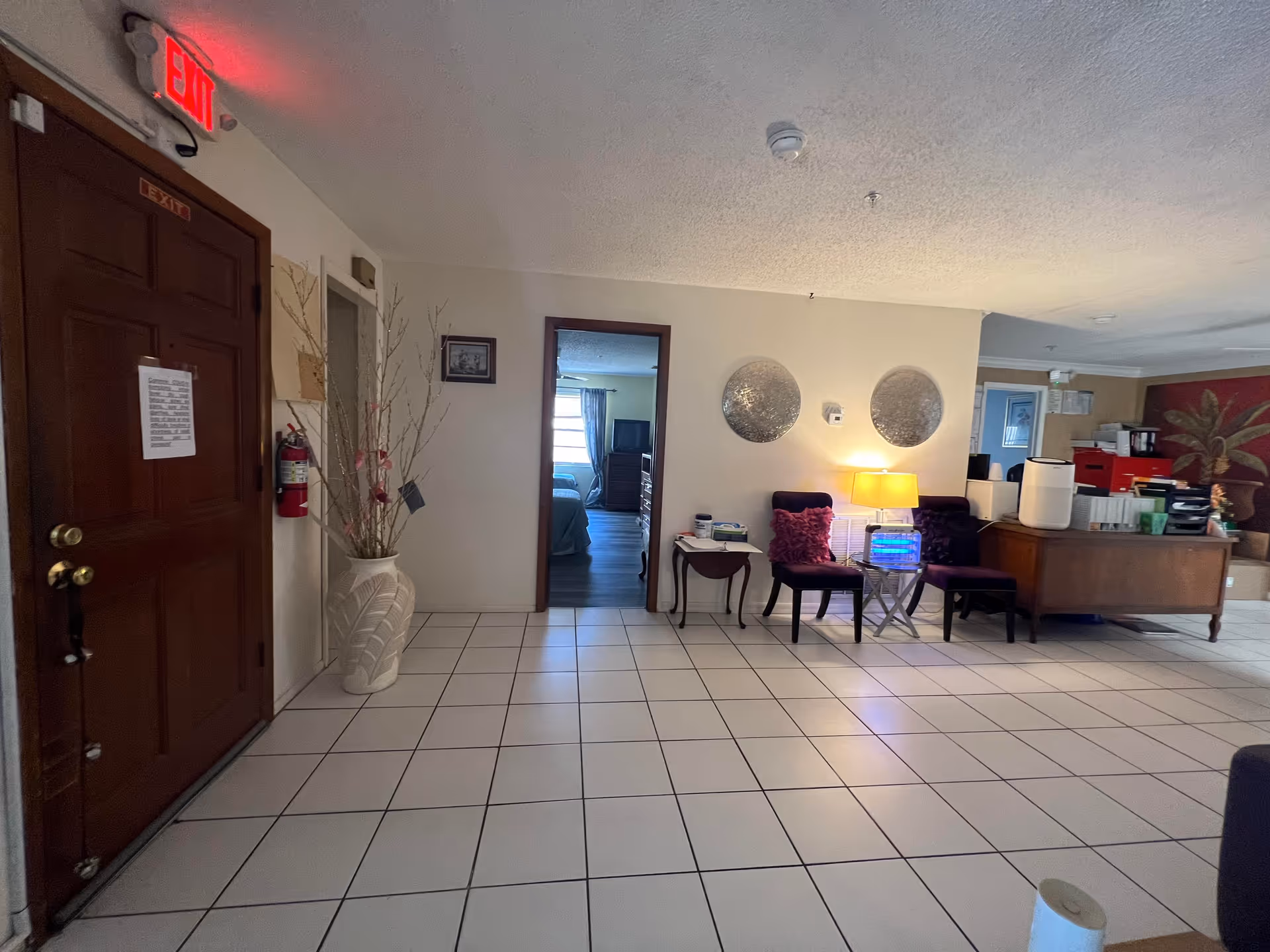 Interior view of a senior living facility showing a tiled floor, a wooden door with an exit sign above it, a large decorative vase with branches, two chairs with purple cushions next to a small table with a lamp, and a desk area with office supplies and a palm tree wall decoration in the background. A doorway leads to a bedroom with a bed and window visible.